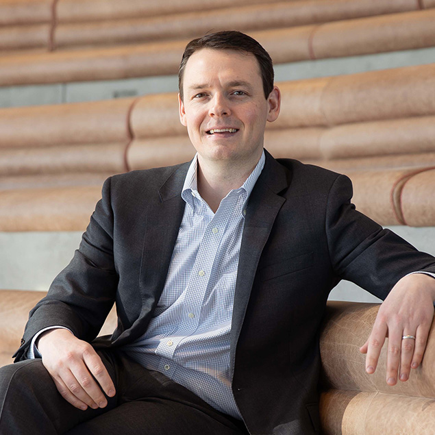 A man in a dark suit and light blue shirt sits on tan bleacher-style steps, smiling with one arm resting on the back of the seat. The background is made of curved wooden seating.