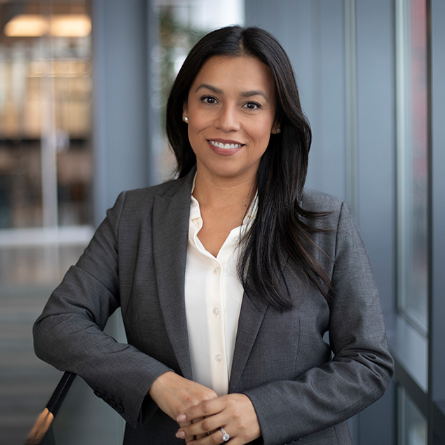 A woman with long dark hair, wearing a gray blazer and white blouse, stands indoors by a glass wall, smiling confidently at the camera.