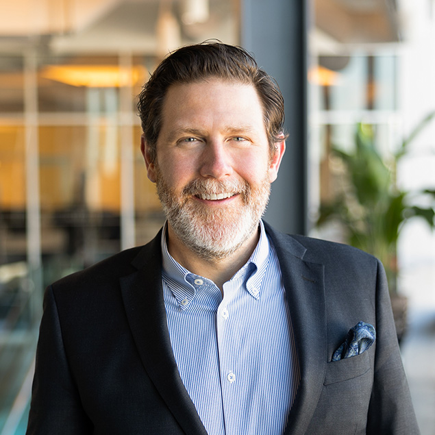 A man with short brown hair and a beard, wearing a dark blazer over a blue striped shirt, smiles while standing indoors near large windows and a potted plant.