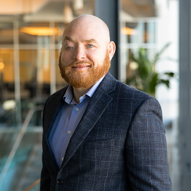 A bald man with a red beard, wearing a blue dress shirt and a dark plaid blazer, stands indoors in front of large windows and a blurred office background, smiling at the camera.