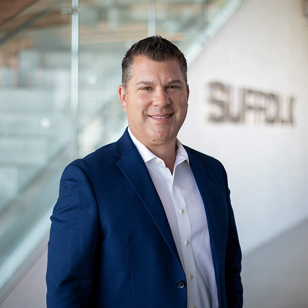 A man in a blue suit and white shirt stands indoors, smiling at the camera. The background features glass railings and the blurred word Suffolk on the wall.