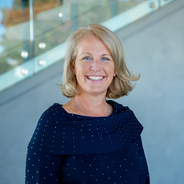 A smiling woman with blonde hair, showcasing executive leadership, wears a navy blue dotted top as she stands indoors in front of a glass railing and gray staircase.