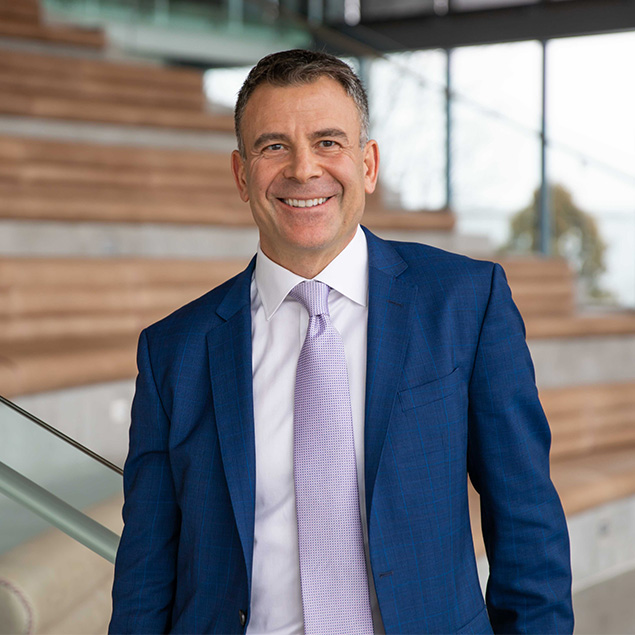 A man in a blue suit and light purple tie smiles while standing indoors near wooden steps and a glass railing.