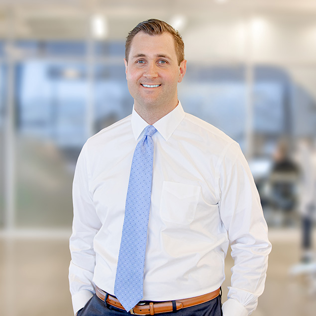 A man stands indoors wearing a white dress shirt, light blue patterned tie, and brown belt, smiling at the camera. The background is a blurred modern office setting.