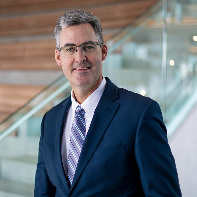 A man in a navy blue suit, white shirt, and striped tie stands indoors near a glass railing and wooden stairs, smiling at the camera and exuding executive leadership.