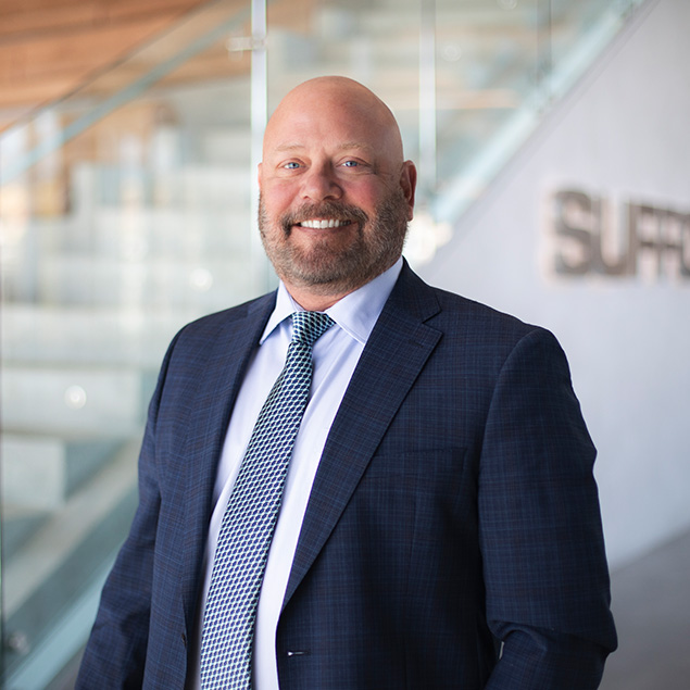 A bald man with a beard, wearing a blue suit, white shirt, and patterned tie, stands smiling in a modern office space with glass railings and blurred signage in the background.