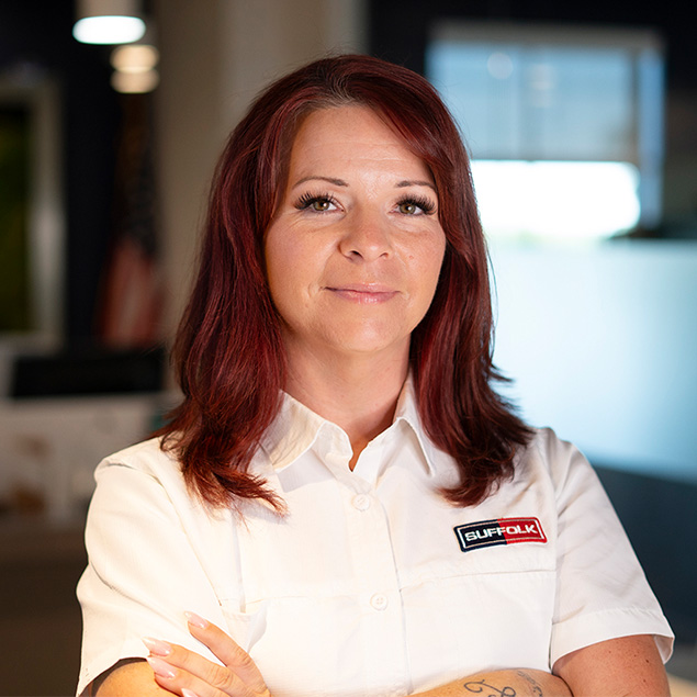 A woman with long red hair, wearing a white short-sleeve button-up shirt with a BUFFOLK patch, stands indoors with arms crossed and smiles confidently at the camera.