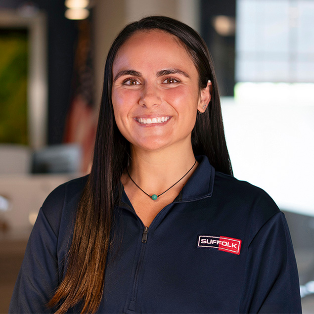 A woman with long dark hair, wearing a navy zip-up shirt with a Suffolk logo, smiles at the camera in a bright, modern office setting.