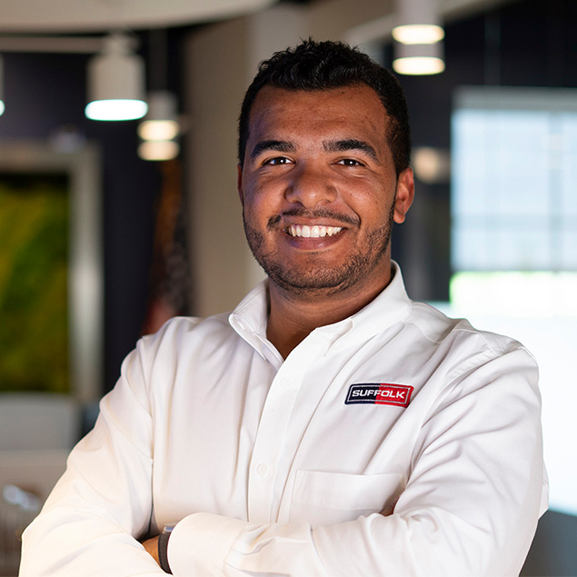 A man with short, dark hair smiles with arms crossed, wearing a white shirt with a Suffolk logo. He stands in a modern, well-lit office space with blurred lights and windows in the background.
