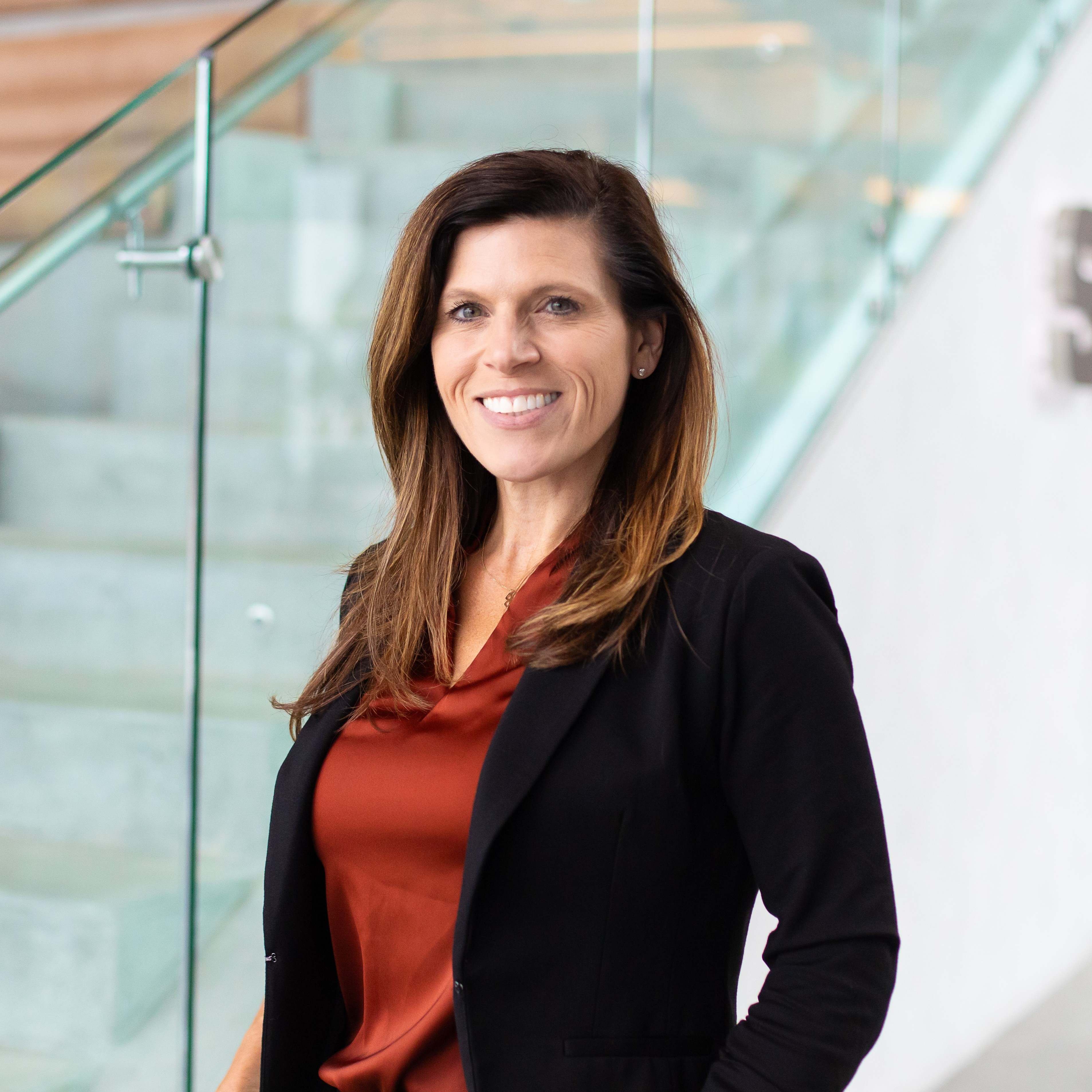 A woman with long brown hair, wearing a black blazer over a rust-colored blouse, stands smiling in front of a modern glass staircase.