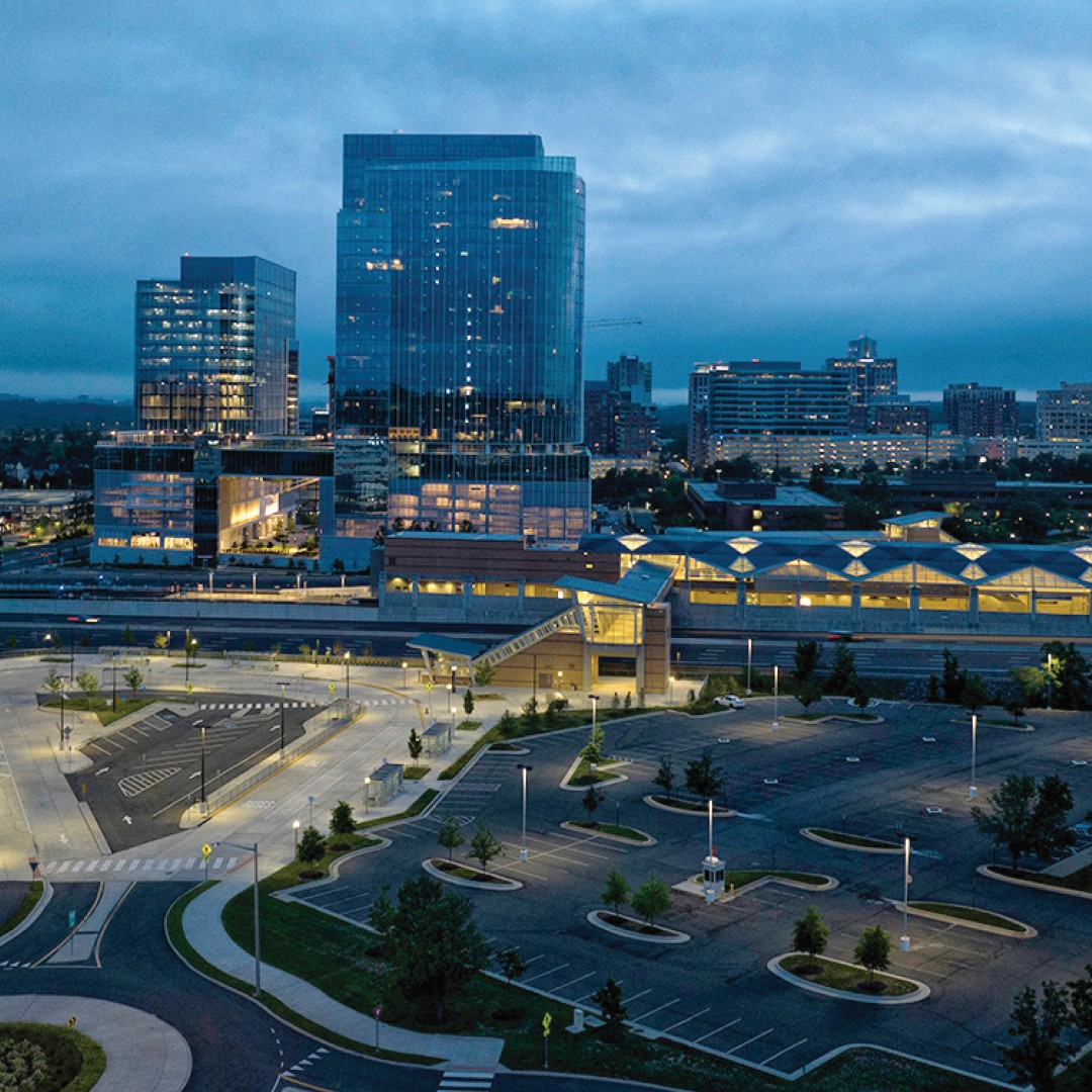 Aerial view of a cityscape at dusk, featuring modern glass buildings, a nearly empty parking lot, illuminated streetlights, and patches of greenery under a cloudy sky.