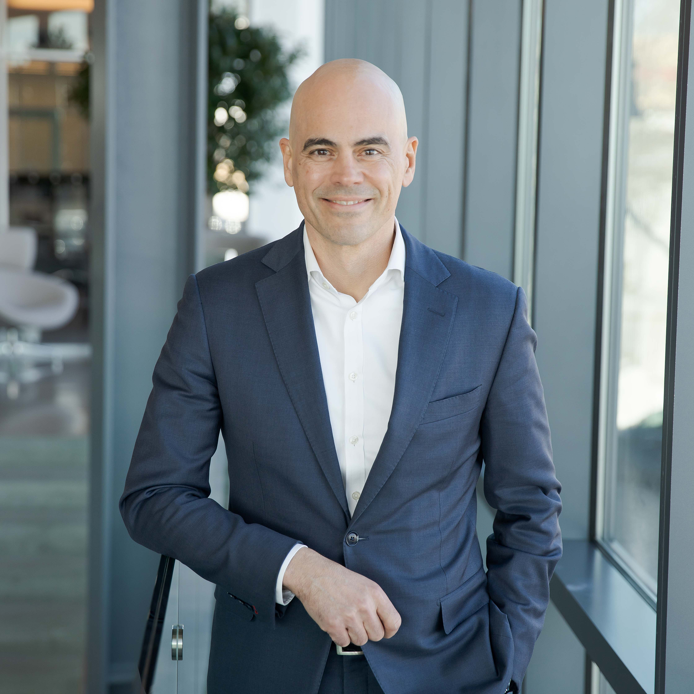 A bald man in a navy blue suit and white shirt stands indoors by large windows, smiling confidently with one hand in his pocket—a modern setting that reflects executive leadership, with touches of greenery visible in the background.