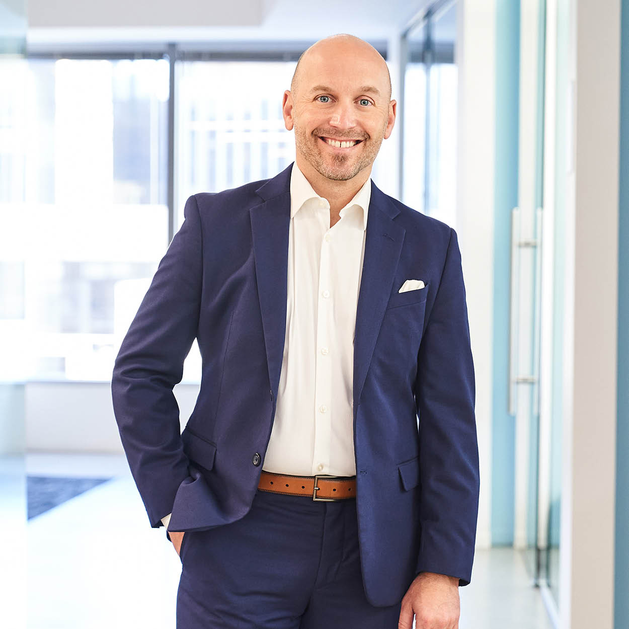 A smiling man in a blue suit and white shirt stands in a bright, modern office hallway with one hand in his pocket.