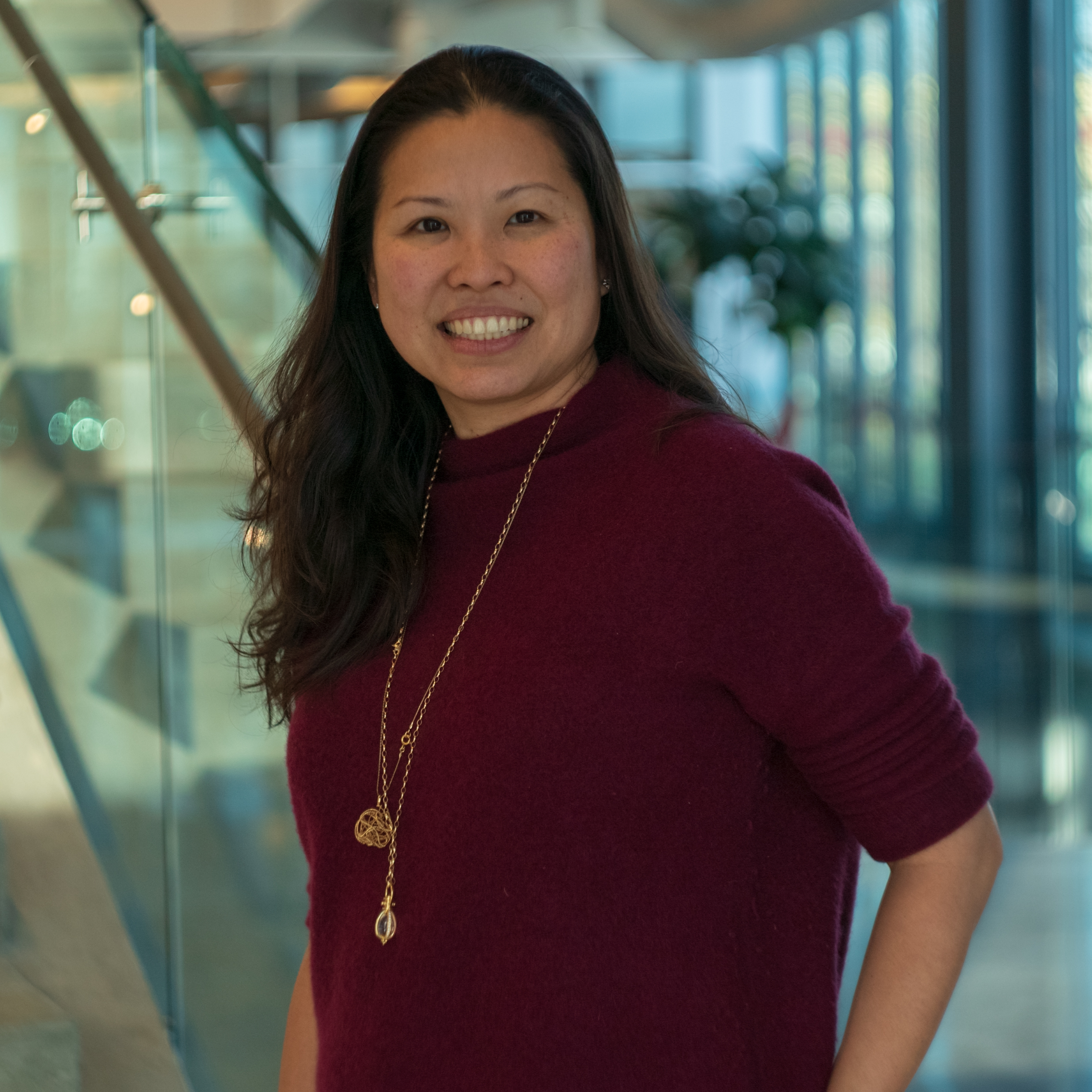 A woman with long dark hair wearing a burgundy sweater and a long gold necklace smiles while standing indoors next to a glass railing, embodying executive leadership. The background includes glass windows and a potted plant.