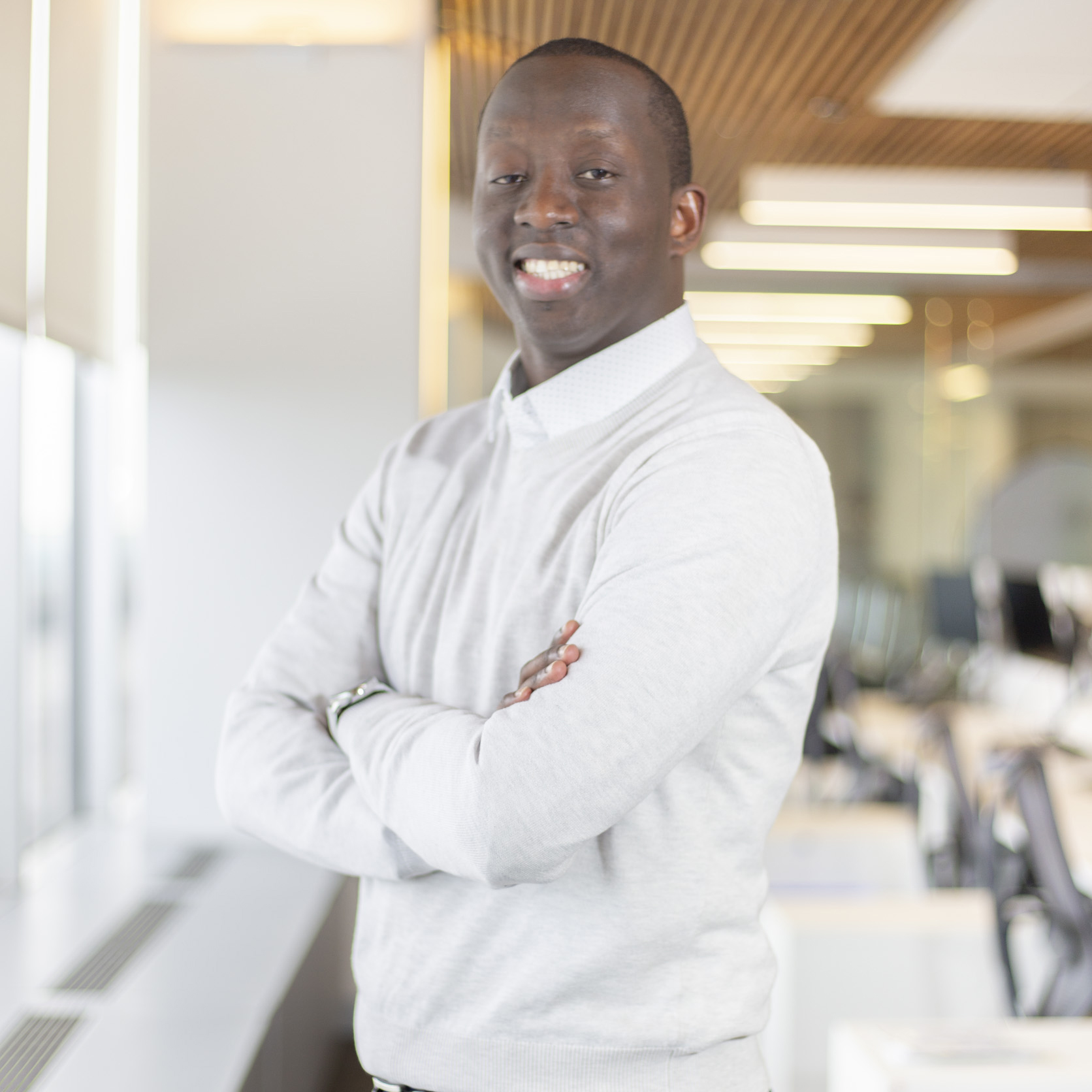 A man wearing a light gray sweater stands confidently with his arms crossed, smiling, in a bright, modern office space with large windows and wooden ceiling panels.