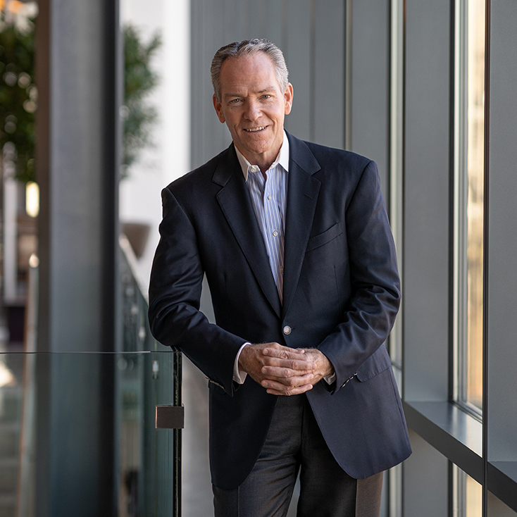 A man in a dark suit and light blue shirt stands indoors, leaning casually against a railing by large windows, smiling at the camera—radiating executive leadership.