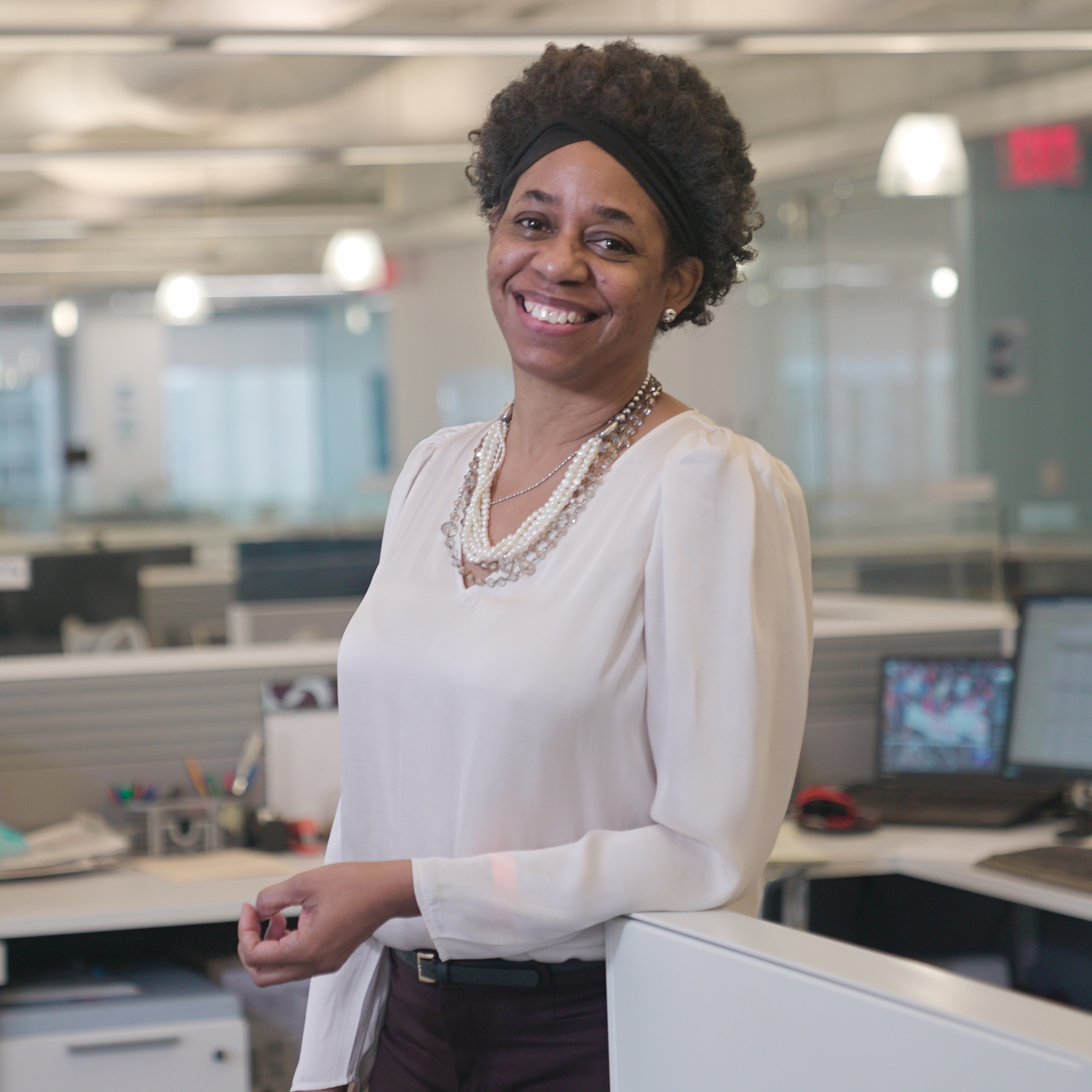 A woman with short curly hair, wearing a white blouse and pearl necklace, smiles while standing in a modern office community with cubicles and computers in the background.