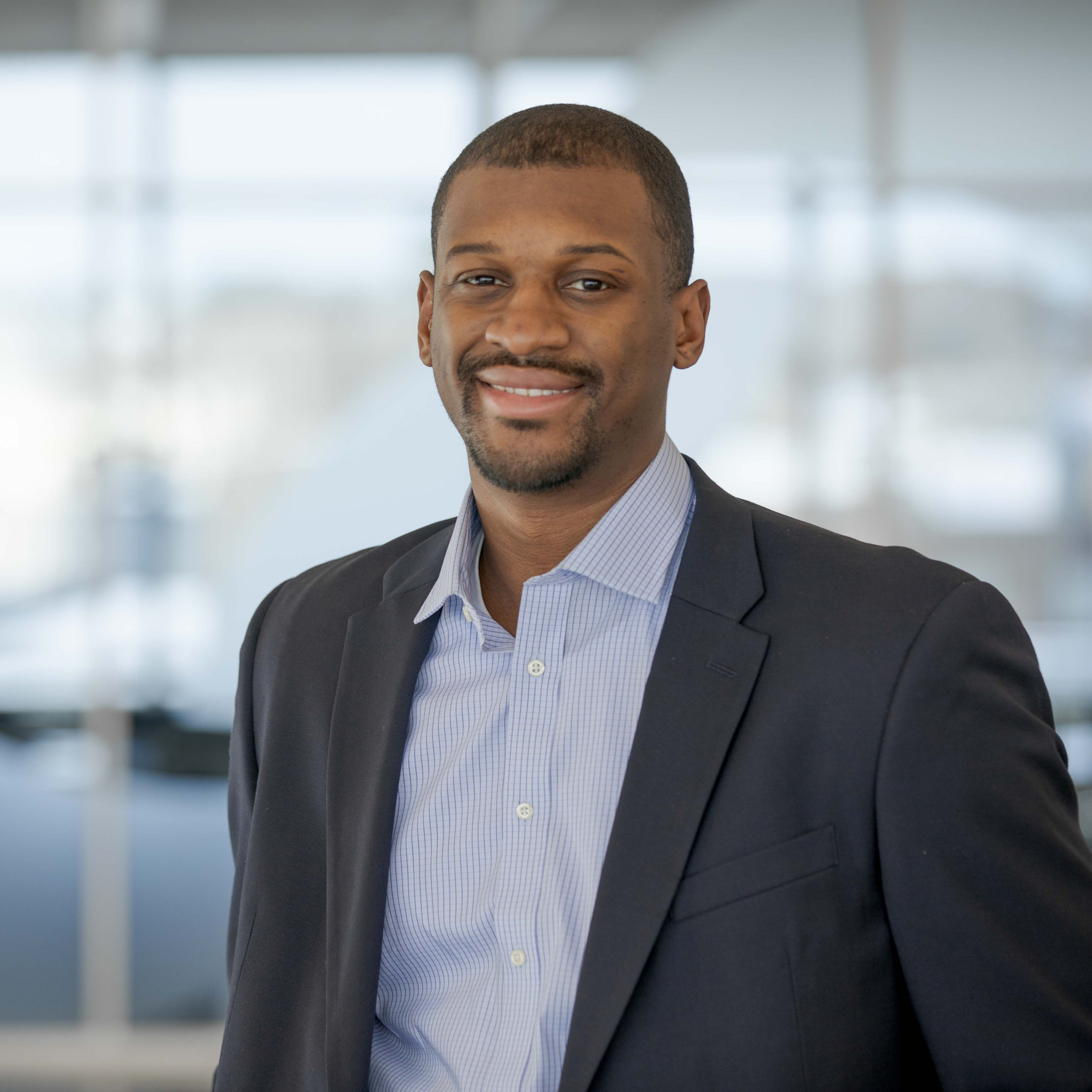 A man wearing a navy blazer and light blue dress shirt smiles at the camera, standing in a modern office with large windows and a blurred background.