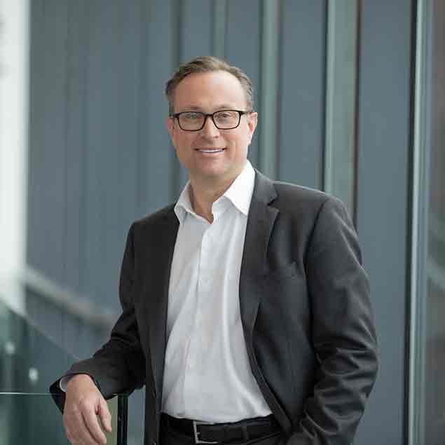 A man wearing glasses, a white dress shirt, and a dark suit jacket stands indoors by a glass railing, smiling at the camera with large windows in the background.