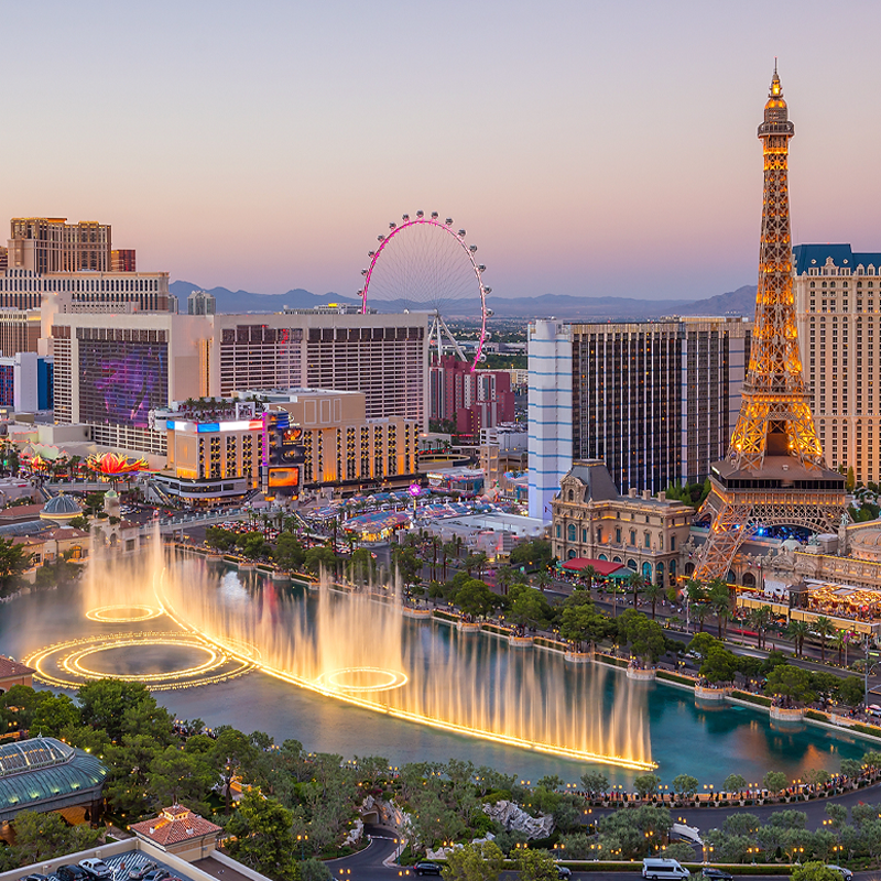Aerial view of the Las Vegas Strip at sunset, featuring the Bellagio fountains performing, the Eiffel Tower replica, colorful hotel buildings, and the High Roller Ferris wheel in the background.
