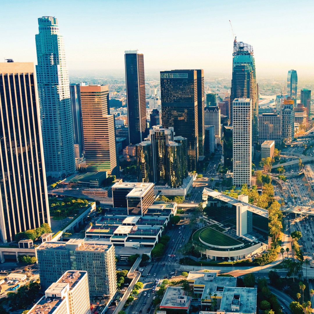 Aerial view of downtown Los Angeles with tall skyscrapers, modern office buildings, and busy city streets bathed in warm sunlight. A construction crane is visible atop a tower under development.