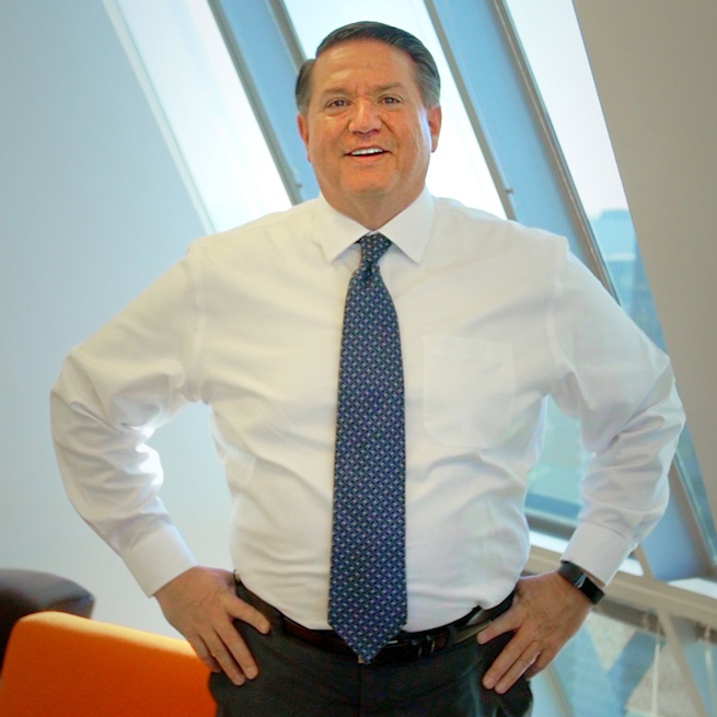 A man in a white dress shirt and patterned blue tie stands smiling with hands on hips in a modern office with large slanted windows and orange and brown furniture in the background.
