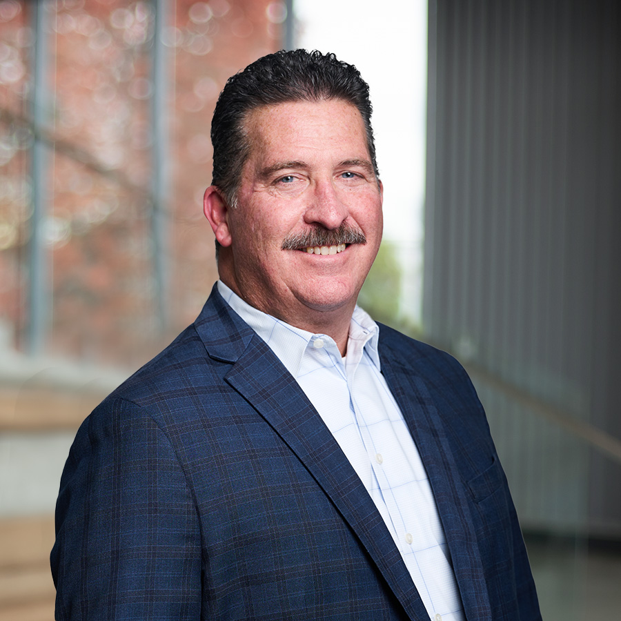 A man with dark hair and a mustache, wearing a blue plaid suit jacket and a light blue shirt, stands indoors smiling, with blurred stairs and large windows in the background.