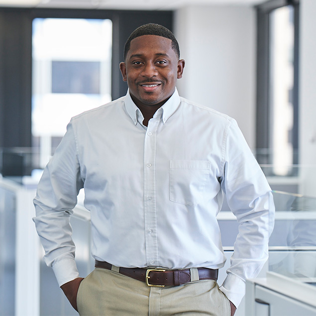 A man in a white dress shirt and khaki pants stands in a modern office, smiling with one hand in his pocket. The office has large windows and white cubicles in the background.