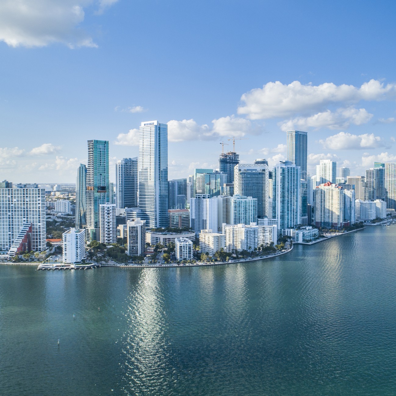Aerial view of a modern city skyline with tall glass skyscrapers and buildings along a curved waterfront under a blue sky with scattered clouds.