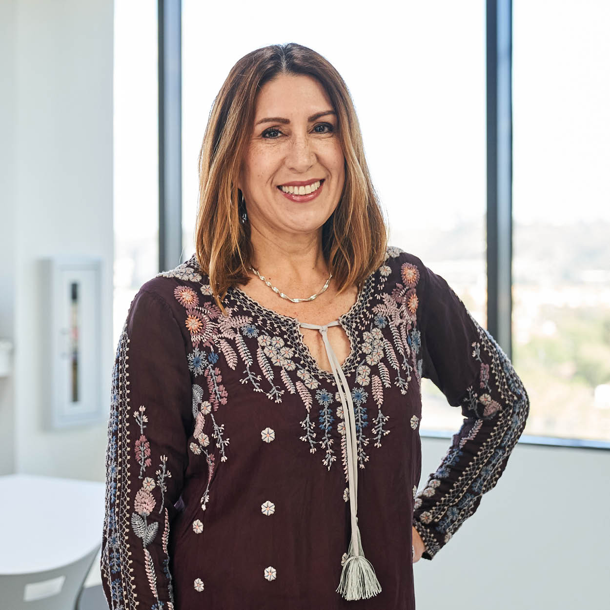 A woman with shoulder-length brown hair, wearing a dark embroidered blouse with tassels, stands smiling in a bright room with large windows in the background.