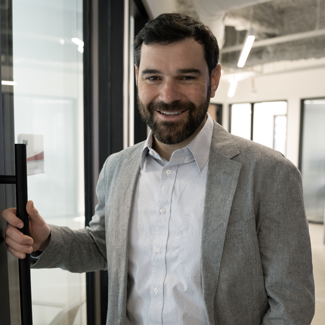 A man with dark hair and a beard, wearing a light gray blazer and white shirt, stands indoors holding a glass door handle and smiling at the camera, ready to join his team in a modern office setting.