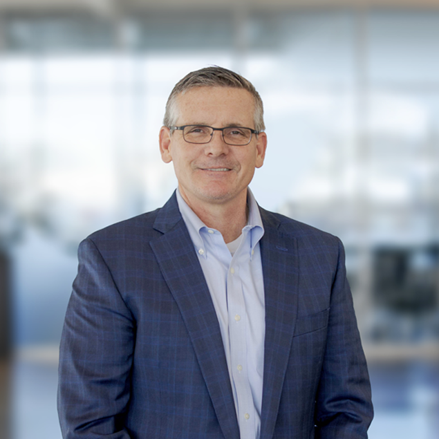 A middle-aged man wearing glasses, a blue plaid suit jacket, and a light blue shirt stands smiling in a modern, blurred office setting, embodying executive leadership.
