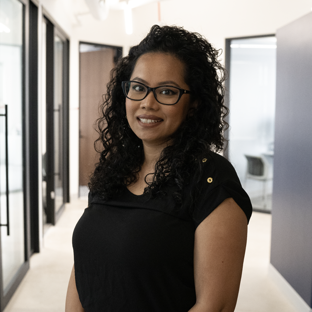 A woman with long curly hair and glasses, wearing a black top, stands smiling in a modern office hallway with glass doors and light-colored walls.