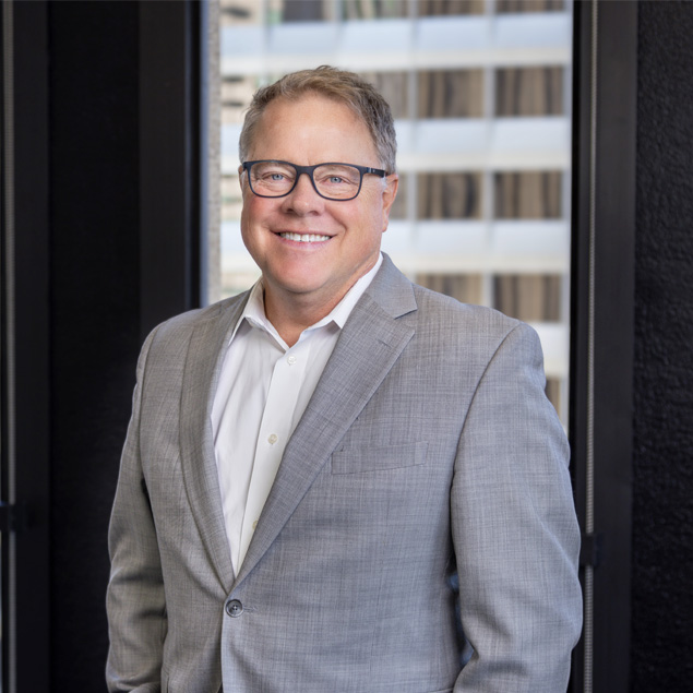 A man wearing glasses and a light gray suit with a white shirt stands indoors, smiling at the camera. Large windows with an urban building view are in the background.