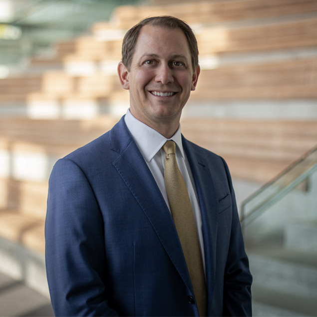 A man in a blue suit, white shirt, and gold tie stands indoors in front of wooden bleacher-style seating, smiling at the camera. The softly blurred background highlights his executive leadership presence.