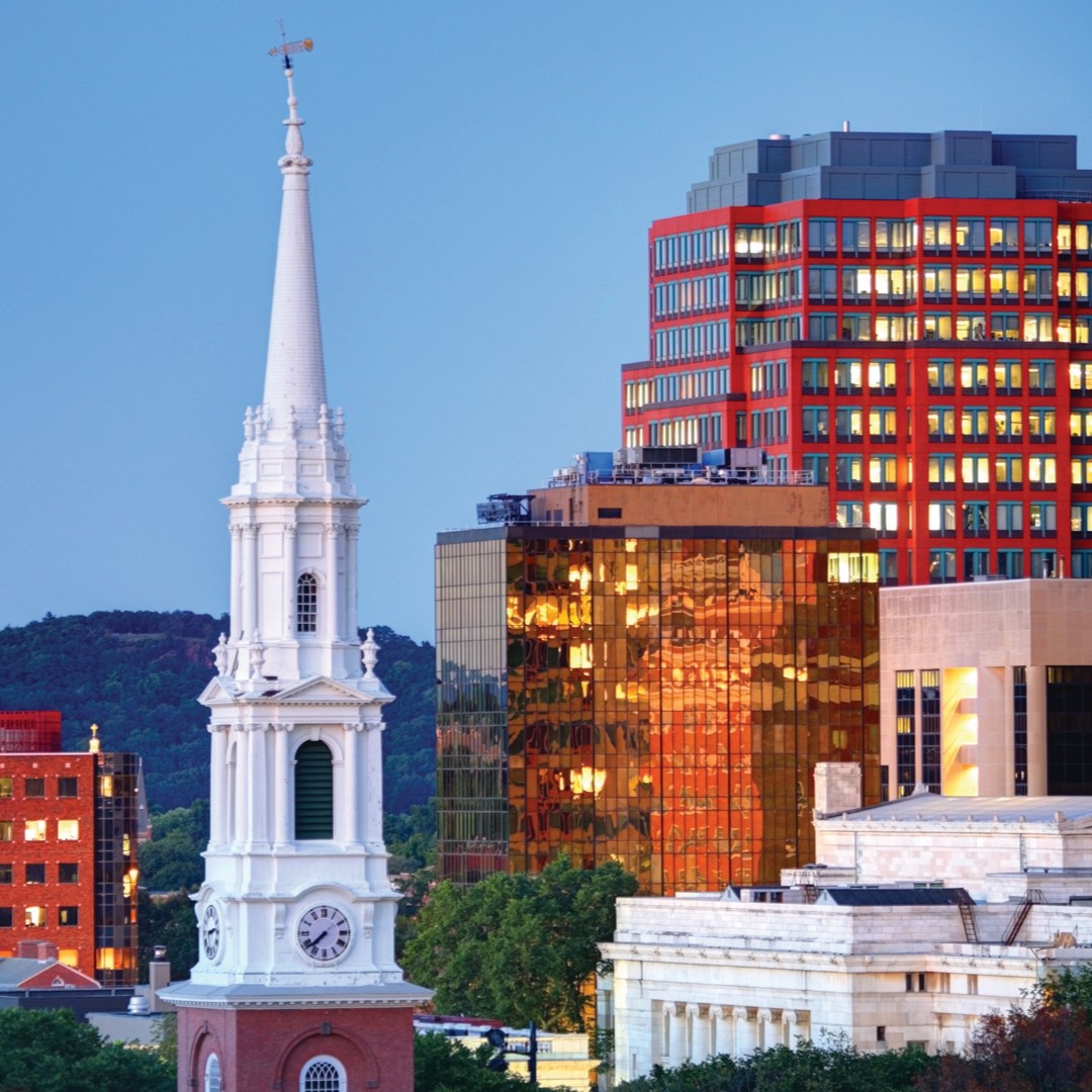 A white church steeple with a clock stands in front of modern glass and red buildings at dusk, with hills and a soft blue sky in the background.