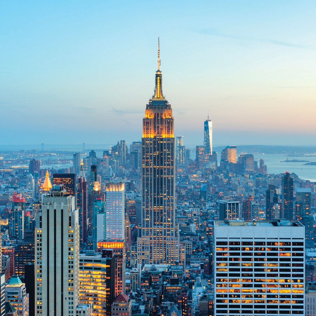 A cityscape of New York City at dusk, featuring the illuminated Empire State Building in the center, surrounded by skyscrapers and buildings, with a soft blue sky in the background.