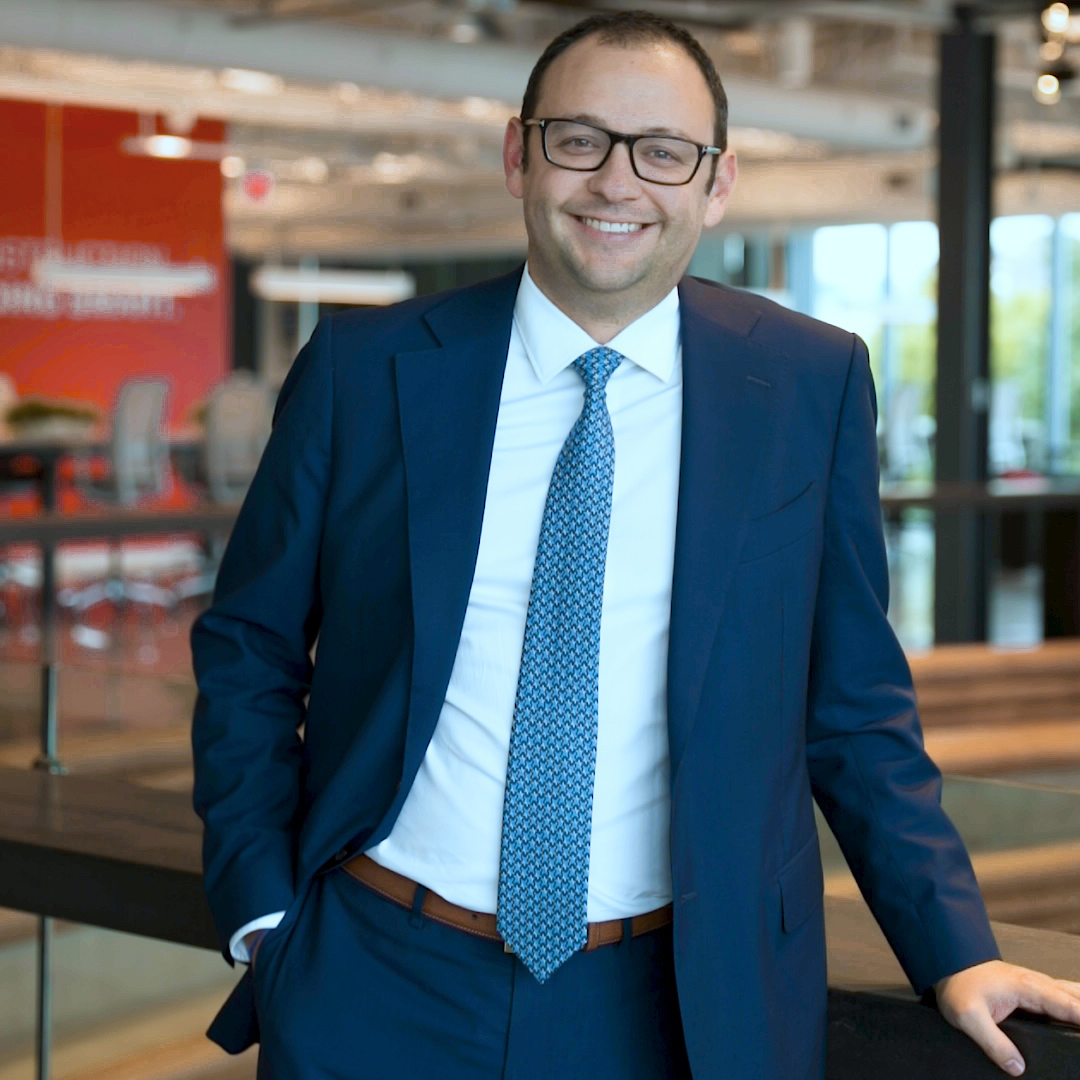 A man wearing glasses, a blue suit, white shirt, and patterned tie stands indoors, smiling with one hand in his pocket. The background features a modern office space with large windows and red accents.