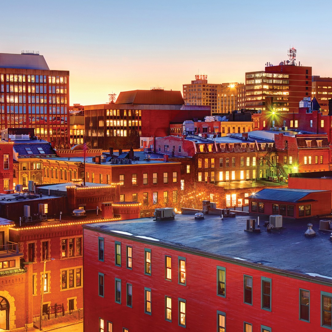 A cityscape at sunset features red brick buildings with illuminated windows and modern office buildings in the background under a clear sky.