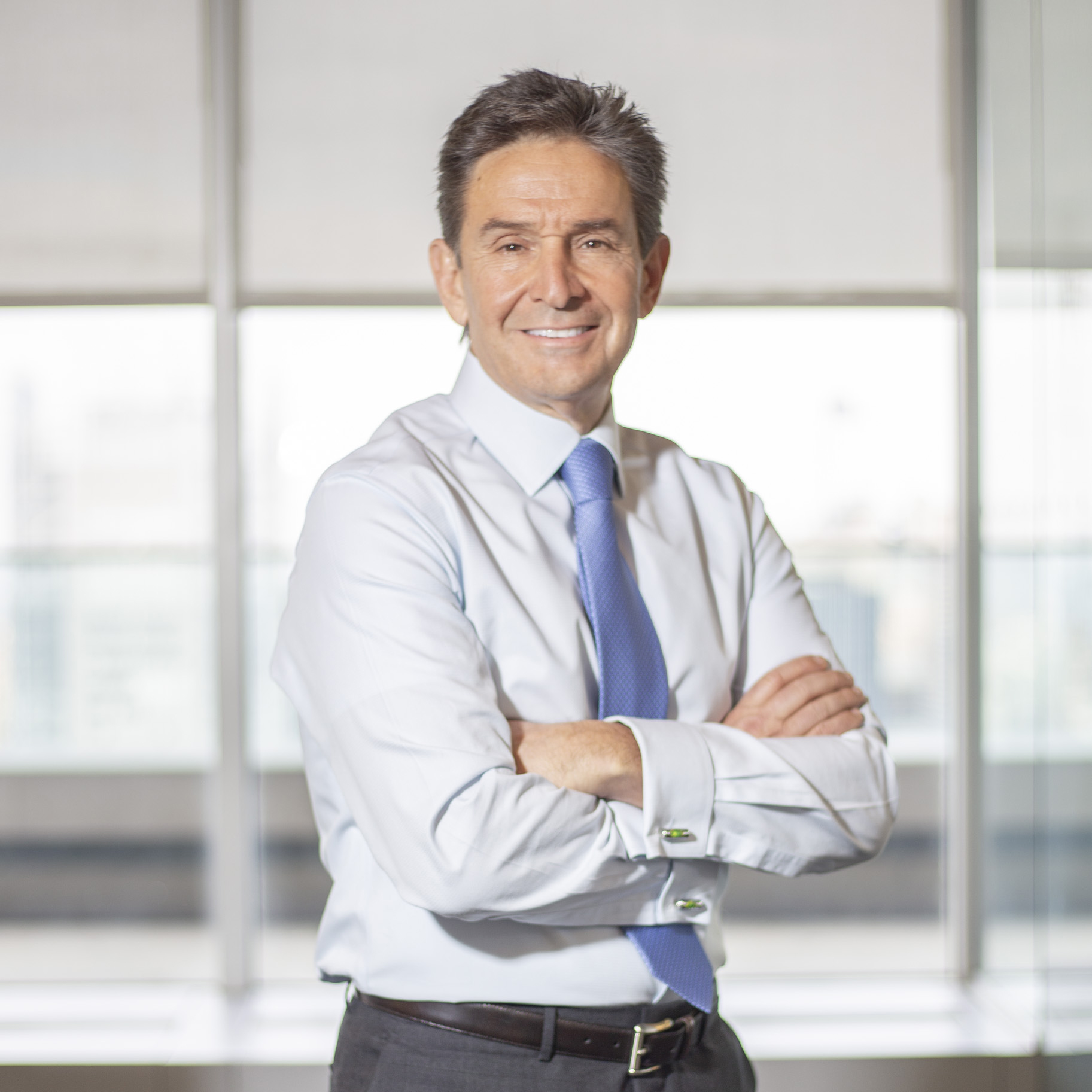 A middle-aged man in a light blue shirt and blue tie stands with arms crossed, smiling, in a bright office with large windows in the background.