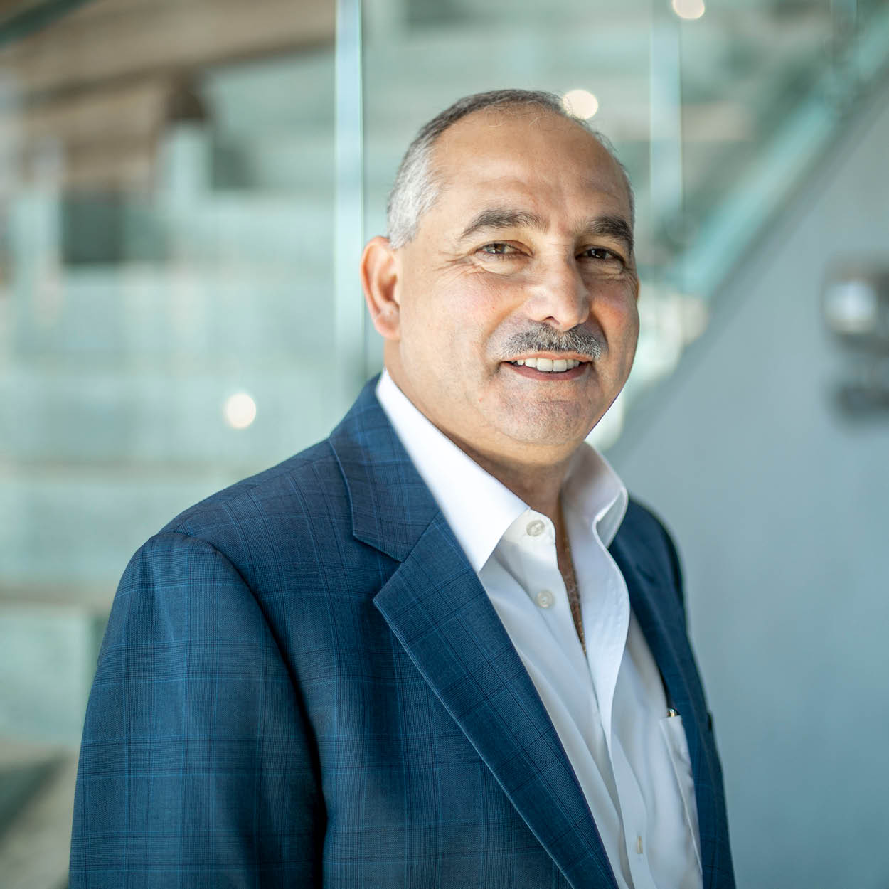 A middle-aged man with short gray hair and a mustache, wearing a blue checked suit jacket and white shirt, smiles while standing indoors in front of a glass staircase.