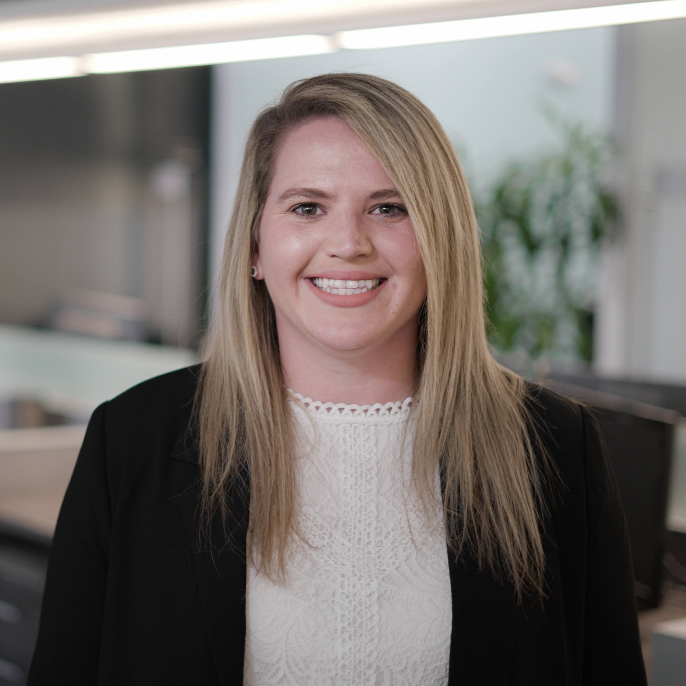 A woman with long blonde hair, wearing a black blazer and white blouse, smiles while standing in a modern office with blurred desks and green plants in the background.