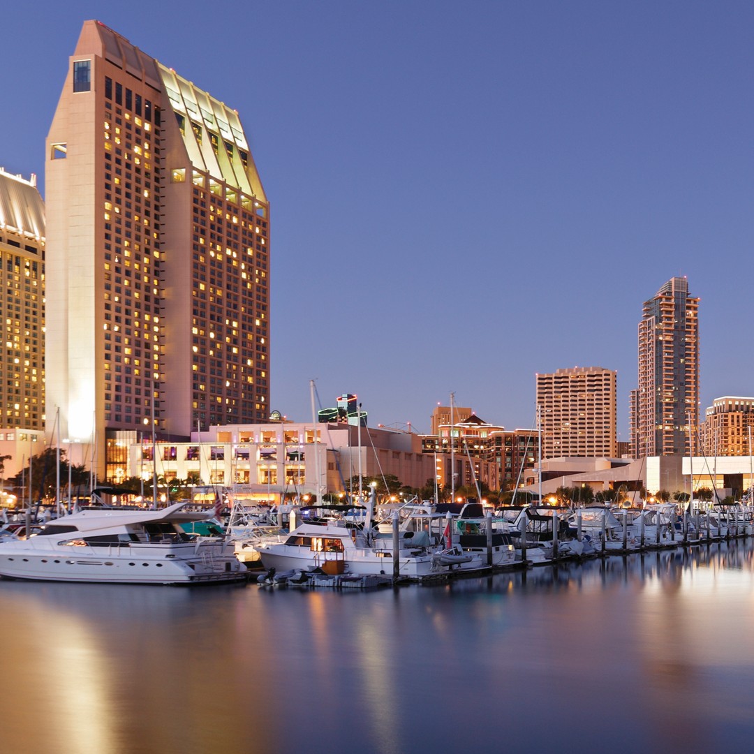 A waterfront marina with docked boats and yachts, tall modern buildings, and a lit-up hotel at dusk, reflecting on calm water under a clear evening sky.