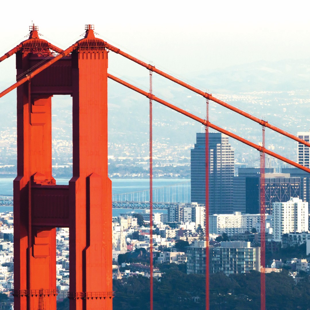 Close-up view of the bright orange Golden Gate Bridge tower with suspension cables, set against a backdrop of downtown San Francisco buildings and a distant bay.