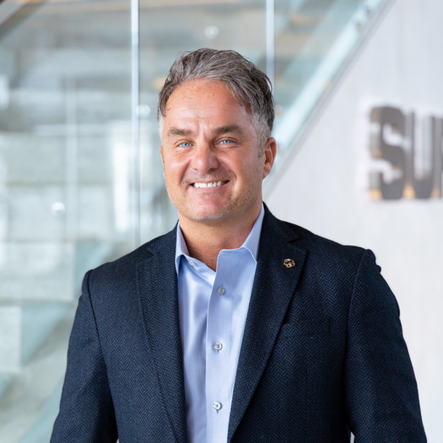 A man with short gray hair in a blue dress shirt and dark blazer smiles while standing indoors in front of a glass staircase and a wall with silver lettering.
