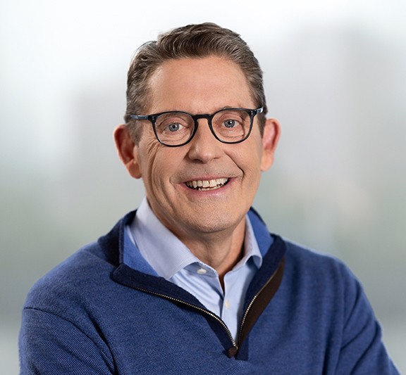 A smiling man with short brown hair and glasses wearing a blue sweater over a light blue collared shirt, with a blurred, neutral background.