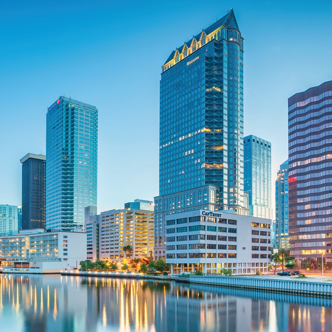City skyline reflected in calm water at dusk, featuring tall modern skyscrapers and office buildings with illuminated windows under a clear blue sky.