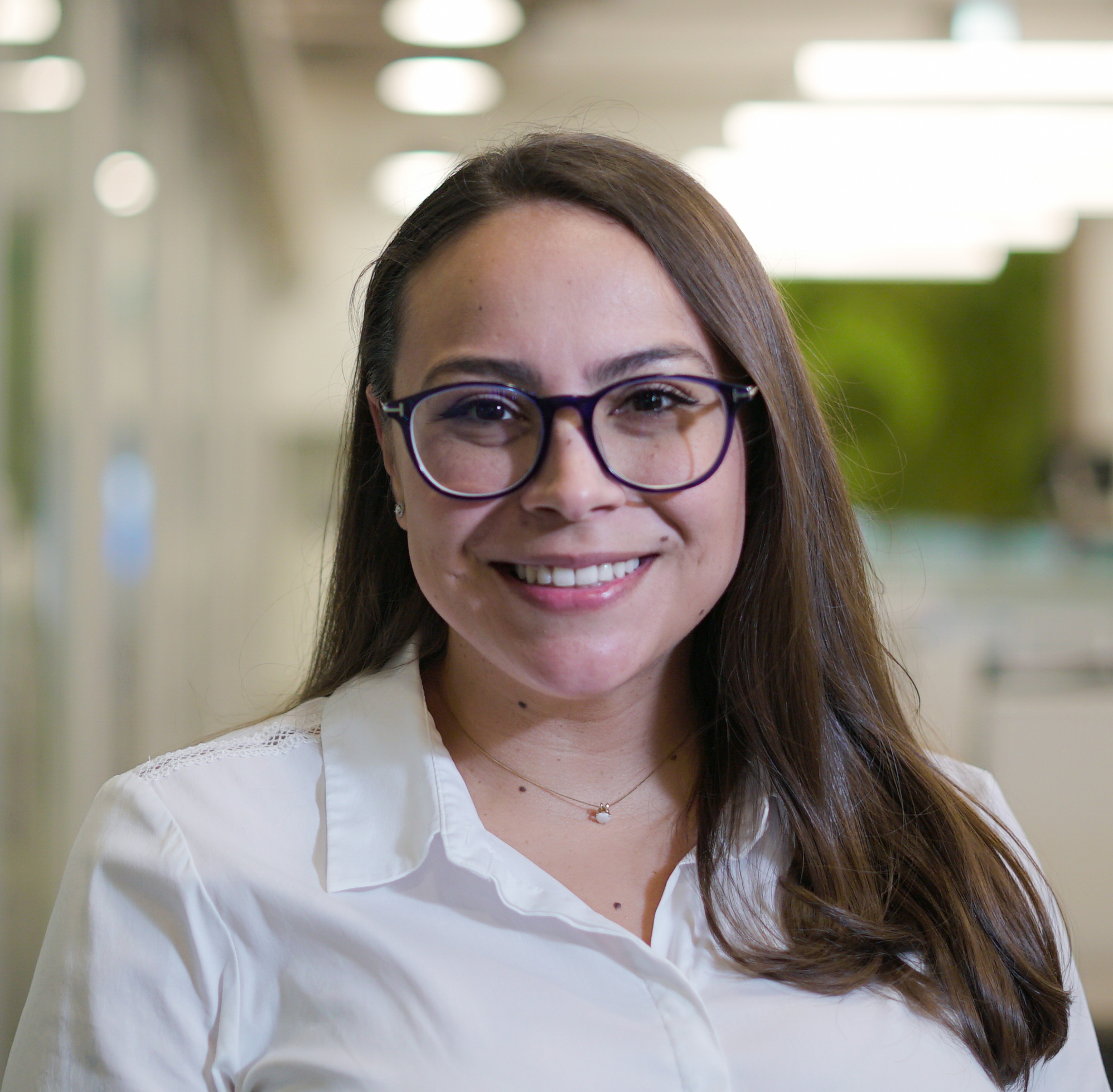 A woman with long brown hair and glasses smiles at the camera. She is wearing a white blouse and a delicate necklace, standing indoors with a blurred office background.