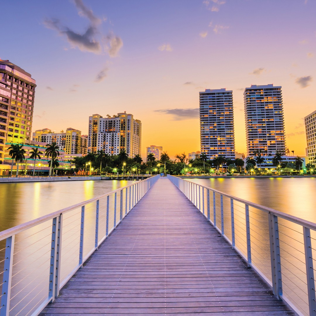 A boardwalk leads over water toward tall, modern buildings and palm trees, illuminated at sunset with a colorful sky in the background.