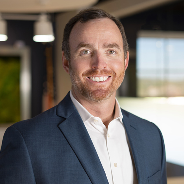 A man with short brown hair and beard, wearing a blue suit jacket and white shirt, smiles while standing in a modern office setting, reflecting confidence as a valued team member amid blurred background elements.